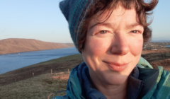 A person wearing a blue jacket and a knitted hat smiles outdoors with a scenic background of a body of water and hilly terrain.