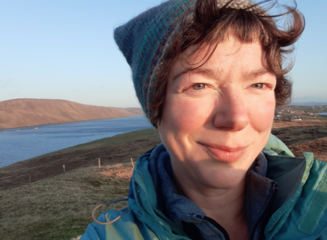 A person wearing a blue jacket and a knitted hat smiles outdoors with a scenic background of a body of water and hilly terrain.