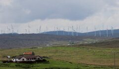 A rural landscape with houses in the foreground and numerous wind turbines spread across the rolling hills in the background under a cloudy sky.