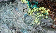 A rock surface with greenish and bluish hues, adorned with strands of yellow and teal plastic, suggesting pollution or human impact on the environment.