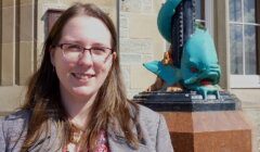 A woman with glasses and long brown hair stands beside a blue dragon sculpture on a brown pedestal outside a building.