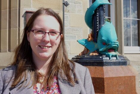 A woman with glasses and long brown hair stands beside a blue dragon sculpture on a brown pedestal outside a building.