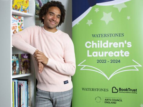 A man in a pink sweater leans against a bookshelf next to a banner that reads "Waterstones Children's Laureate 2022-2024.