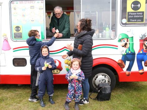 A man serves ice cream from a truck decorated with video game character graphics to a family with three children who are standing outside. A 'Please keep your distance' sign is on the truck.