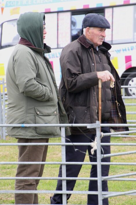 Two individuals in coats stand outside near a metal fence. One holds a wooden cane. An ice cream truck is visible in the background.