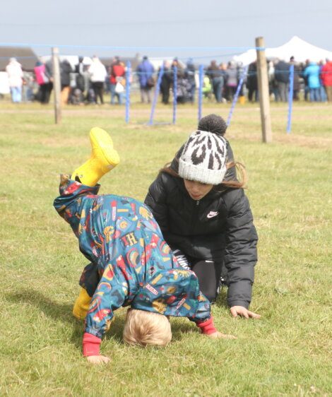 A child wearing a colorful raincoat and yellow boots is doing a headstand on the grass, assisted by another child in a black jacket and beanie. People are gathered in the background.