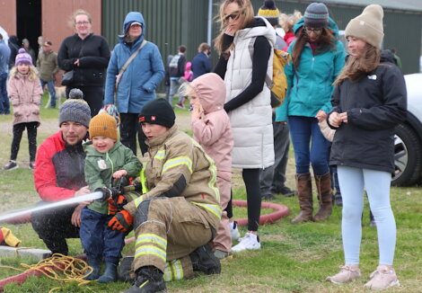 A firefighter helps a young boy hold a fire hose while a group of people, including children and adults, watch the demonstration outdoors on a grassy area.