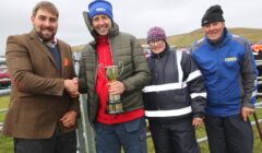 A man in a brown jacket shakes hands with a man in a red jacket holding a trophy. Two other people, one wearing a reflective vest and the other in blue, stand beside them outdoors on a cloudy day.