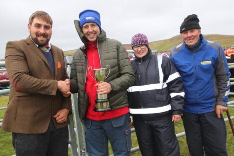 A man in a brown jacket shakes hands with a man in a red jacket holding a trophy. Two other people, one wearing a reflective vest and the other in blue, stand beside them outdoors on a cloudy day.