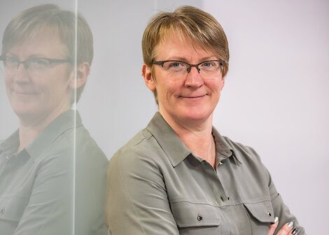 A person with short hair and glasses stands with arms crossed, leaning against a reflective glass surface, wearing a gray button-up shirt.