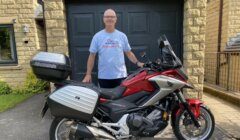 A man stands smiling next to a red Honda motorcycle with side and rear storage boxes. He is wearing a t-shirt with "Royal Air Forces Association" written on it. The setting appears to be a driveway.