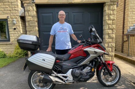 A man stands smiling next to a red Honda motorcycle with side and rear storage boxes. He is wearing a t-shirt with "Royal Air Forces Association" written on it. The setting appears to be a driveway.