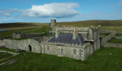 A stone building with crenellations and a central tower is surrounded by scaffolding, indicating restoration work. It sits in a large, grassy, rural area under a blue sky with clouds.