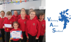 A group of children wearing red hoodies smile while holding a certificate. The Voluntary Action Shetland logo is displayed to the right of the group.