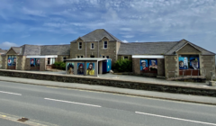 A stone building with mural paintings of various figures on its exterior walls, a small central covered area, and a two-lane road in the foreground under a partly cloudy sky.