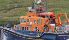 Lifeboat with orange hull and multiple crew members in safety gear operating in a coastal area with grassy hills in the background.