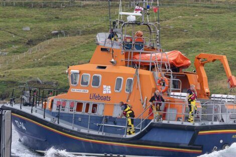 Lifeboat with orange hull and multiple crew members in safety gear operating in a coastal area with grassy hills in the background.