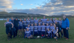 A rugby team poses together on a field after a game, smiling and wearing matching jerseys. Hilly terrain and a partly cloudy sky are visible in the background.