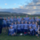 A rugby team poses together on a field after a game, smiling and wearing matching jerseys. Hilly terrain and a partly cloudy sky are visible in the background.