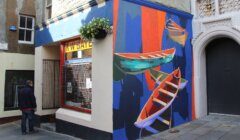 A man stands outside a building corner adorned with a colorful mural of boats. The building has a window with a yellow sign above it and a hanging flower basket on the wall.