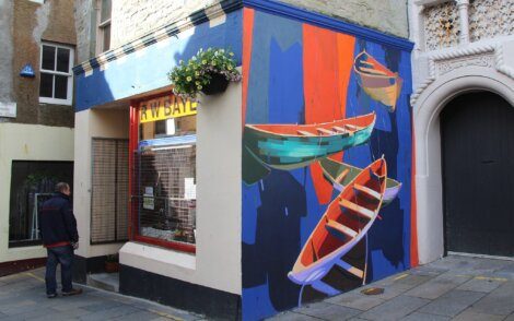 A man stands outside a building corner adorned with a colorful mural of boats. The building has a window with a yellow sign above it and a hanging flower basket on the wall.