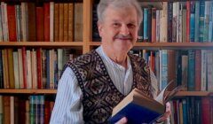 An elderly man with gray hair and a mustache stands smiling in front of a bookshelf, holding an open book. He wears a patterned sweater vest over a button-up shirt.