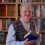 An elderly man with gray hair and a mustache stands smiling in front of a bookshelf, holding an open book. He wears a patterned sweater vest over a button-up shirt.