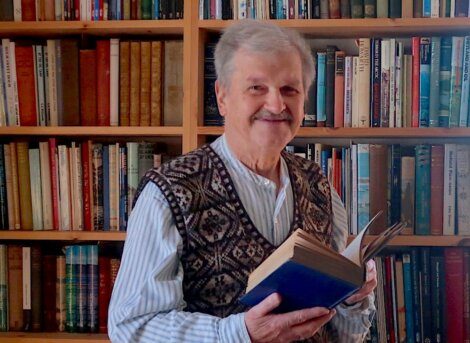 An elderly man with gray hair and a mustache stands smiling in front of a bookshelf, holding an open book. He wears a patterned sweater vest over a button-up shirt.