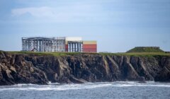 A large industrial building stands on a grassy cliff’s edge overlooking the ocean, with waves crashing against the rocky shoreline below.