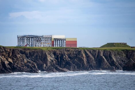 A large industrial building stands on a grassy cliff’s edge overlooking the ocean, with waves crashing against the rocky shoreline below.