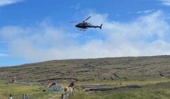 A helicopter hovers above a grassy hill, transporting materials with a long rope. Construction workers and equipment are visible below, with a dirt path leading towards the site.