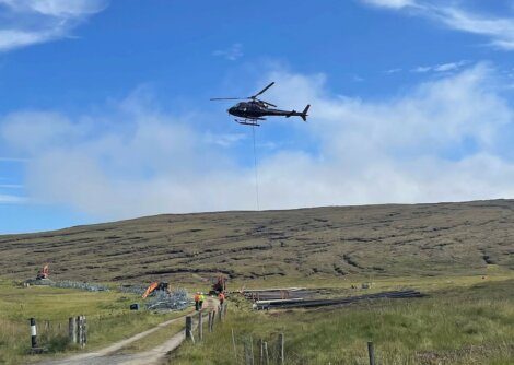 A helicopter hovers above a grassy hill, transporting materials with a long rope. Construction workers and equipment are visible below, with a dirt path leading towards the site.