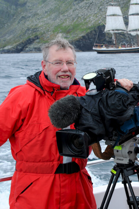 A man in a red jacket stands on a boat with a professional video camera. A sailing ship with white sails is visible in the background on a body of water with rocky hillsides.