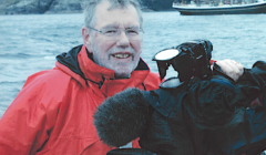 A person in a red jacket stands outdoors near water, holding a large video camera. A boat is visible in the background.