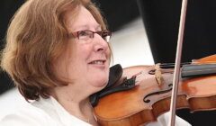 A person with short brown hair and glasses is playing a violin and smiling. They are wearing a white shirt.