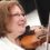 A person with short brown hair and glasses is playing a violin and smiling. They are wearing a white shirt.