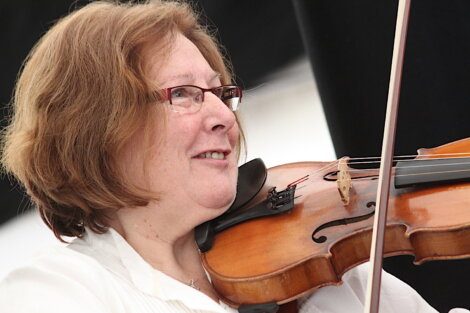 A person with short brown hair and glasses is playing a violin and smiling. They are wearing a white shirt.