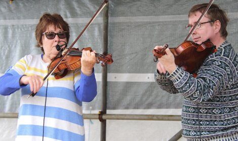 Two people playing violins, one wearing sunglasses and a striped sweater, and the other wearing glasses and a patterned sweater. They are performing on an outdoor stage with a metal mesh background.