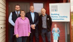 Four people stand in front of a building next to a sign that reads "Building Homes, Creating Communities," associated with the Highland Housing Association.