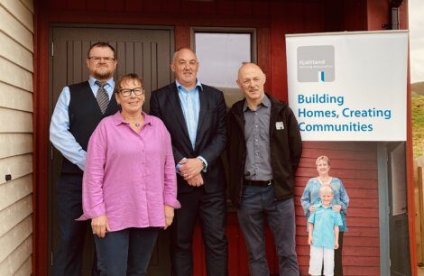 Four people stand in front of a building next to a sign that reads "Building Homes, Creating Communities," associated with the Highland Housing Association.