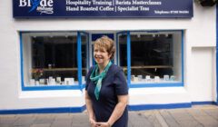 A person stands in front of a shop with a sign reading "Blòths Welcome," advertising hospitality training, barista masterclasses, hand-roasted coffee, and specialist teas.