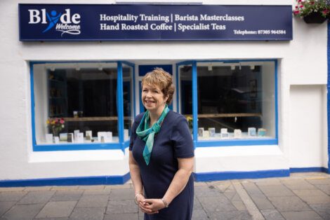 A person stands in front of a shop with a sign reading "Blòths Welcome," advertising hospitality training, barista masterclasses, hand-roasted coffee, and specialist teas.