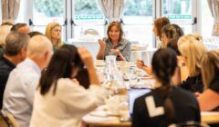 A group of people sit around a table engaged in discussion during a meeting; one woman is speaking while others listen attentively.