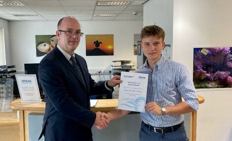 Two men shaking hands in an office; one holds a "Certificate of Scholarship" with the "Ocean Futures" logo.