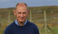 A middle-aged man with a bald head and a slight smile wearing a blue sweater stands outdoors in a grassy rural area with wooden fences in the background.