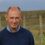 A middle-aged man with a bald head and a slight smile wearing a blue sweater stands outdoors in a grassy rural area with wooden fences in the background.