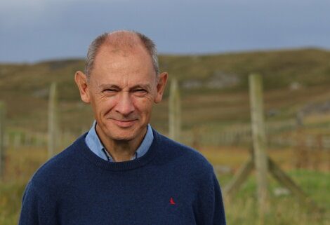 A middle-aged man with a bald head and a slight smile wearing a blue sweater stands outdoors in a grassy rural area with wooden fences in the background.