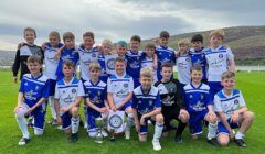 A group of young boys in blue and white football uniforms pose on a grassy field with two trophies.