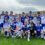 A group of young boys in blue and white football uniforms pose on a grassy field with two trophies.