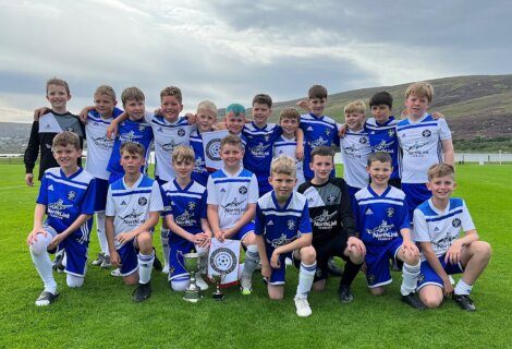 A group of young boys in blue and white football uniforms pose on a grassy field with two trophies.